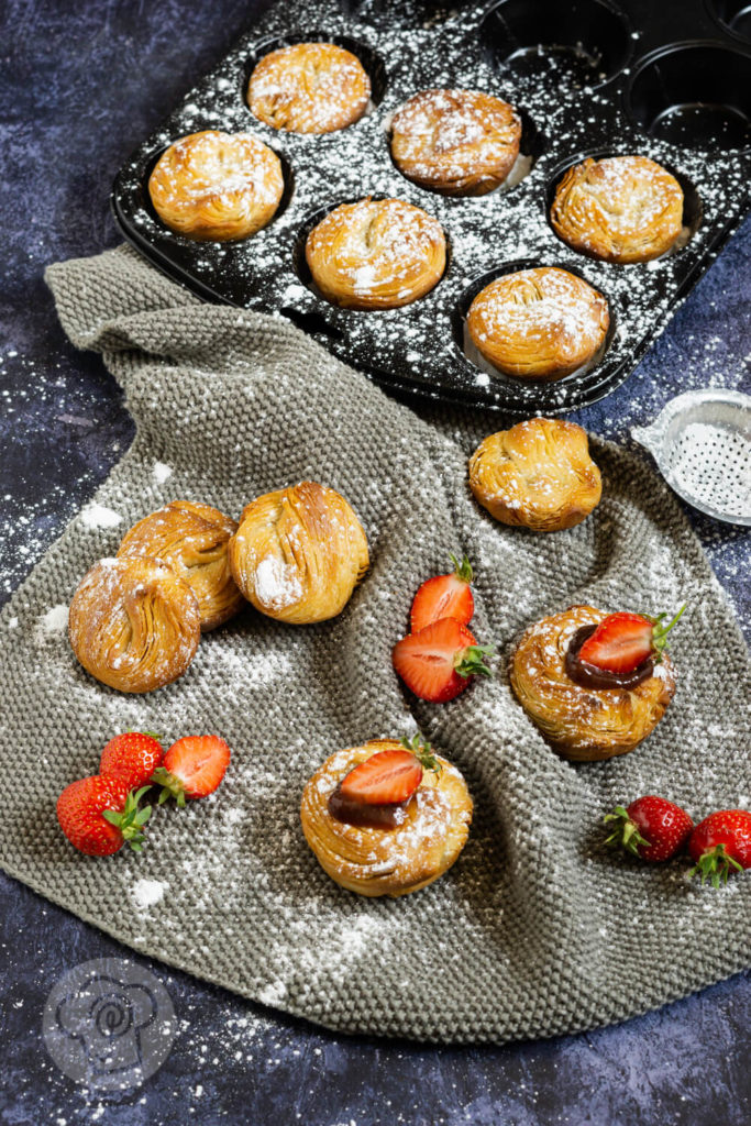 Cruffins von oben fotorgafiert, teilweise im Muffinblech, teilweise auf einem grauen Tuch