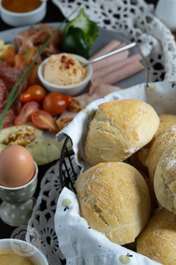 Einfache Frühstücksbrötchen im Brotkorb mit Brot und Grissini. Dazu eine Wurstplatte, Eier und Kaffee.