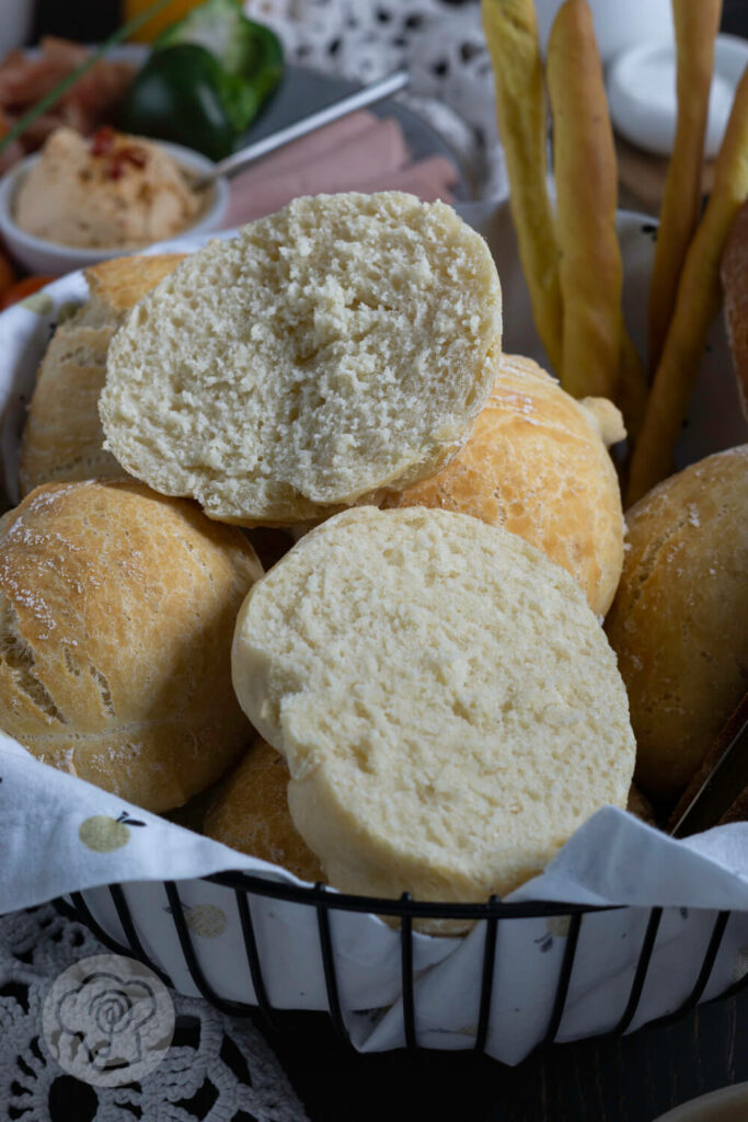 Einfache Frühstücksbrötchen im Brotkorb aufgeschnitten mit Brot und Grissini. Dazu eine Wurstplatte im Hintergrund.