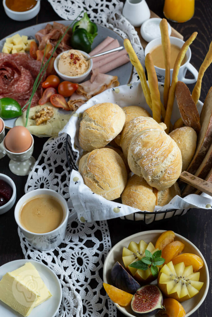 Frühstücksbrötchen im Brotkorb mit Brot und Grissini. Dazu eine Wurstplatte, Eier und Kaffee.