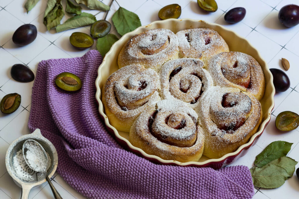 Hefeteigschnecken mit Pflaumen in der Form. Dazu aufgeschnittene Früchte, Blätter und Puderzucker.