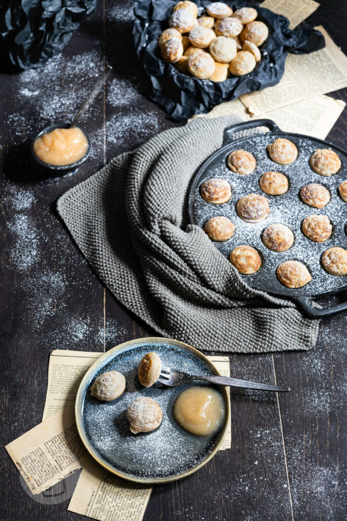 Poffertjes auf einem Teller mit Puderzucker und Apfelbrei. Pfanne mit Poffertjes im Hintergrund.