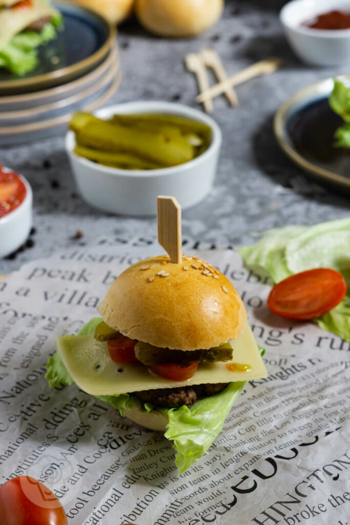 Mini Burger. Im Hintergrund Soße, Teller, Tomaten und Gurken.