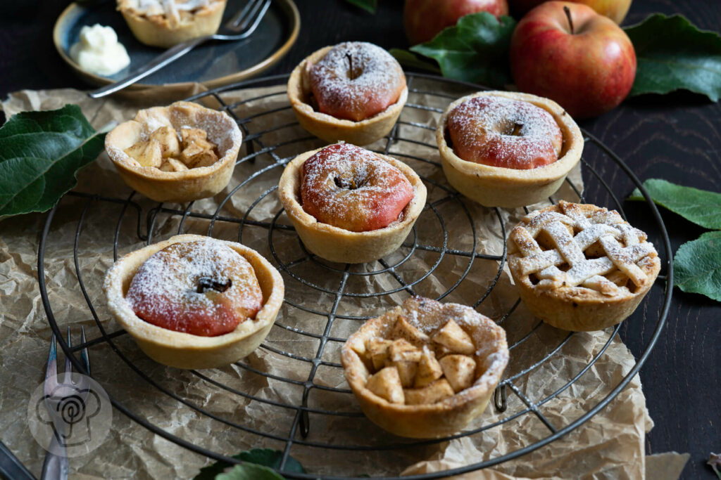 Mini Apple Pies auf einem Kuchenrost. Äpfel im Hintergrund.