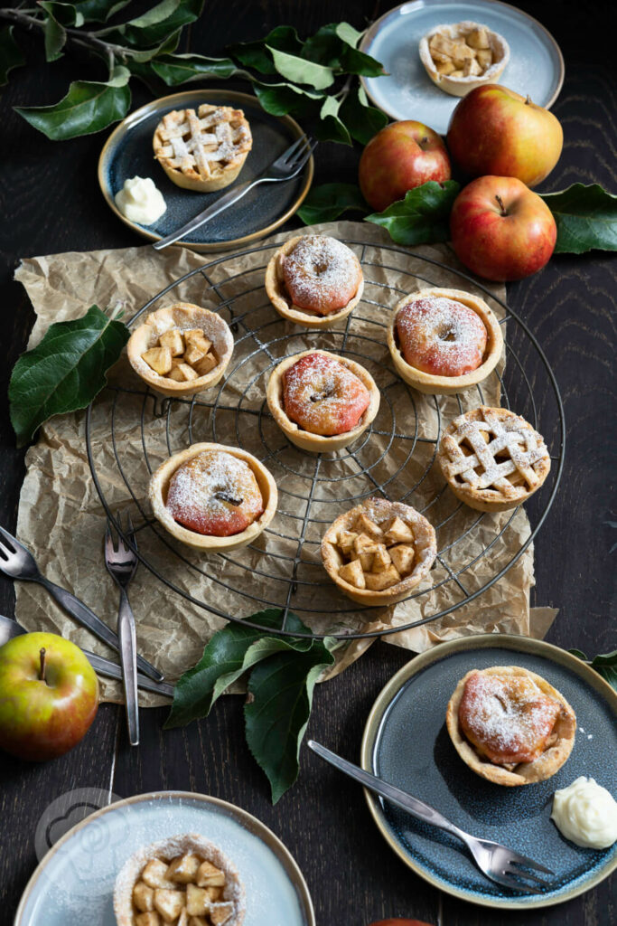 Mini Apple Pies auf einem Kuchenrost und auf Tellern. Äpfel im Hintergrund.