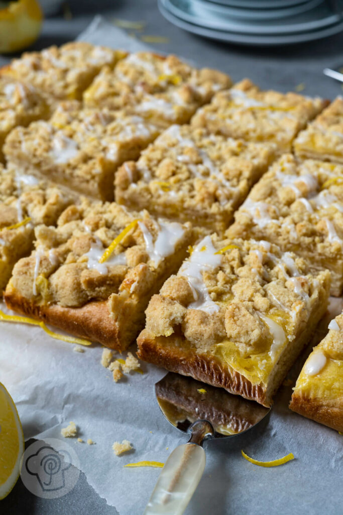 Zitronen Streuselkuchen in Stücke geschnitten mit einer Kuchenschaufel. Geschirr und Zitronen im Hintergrund.