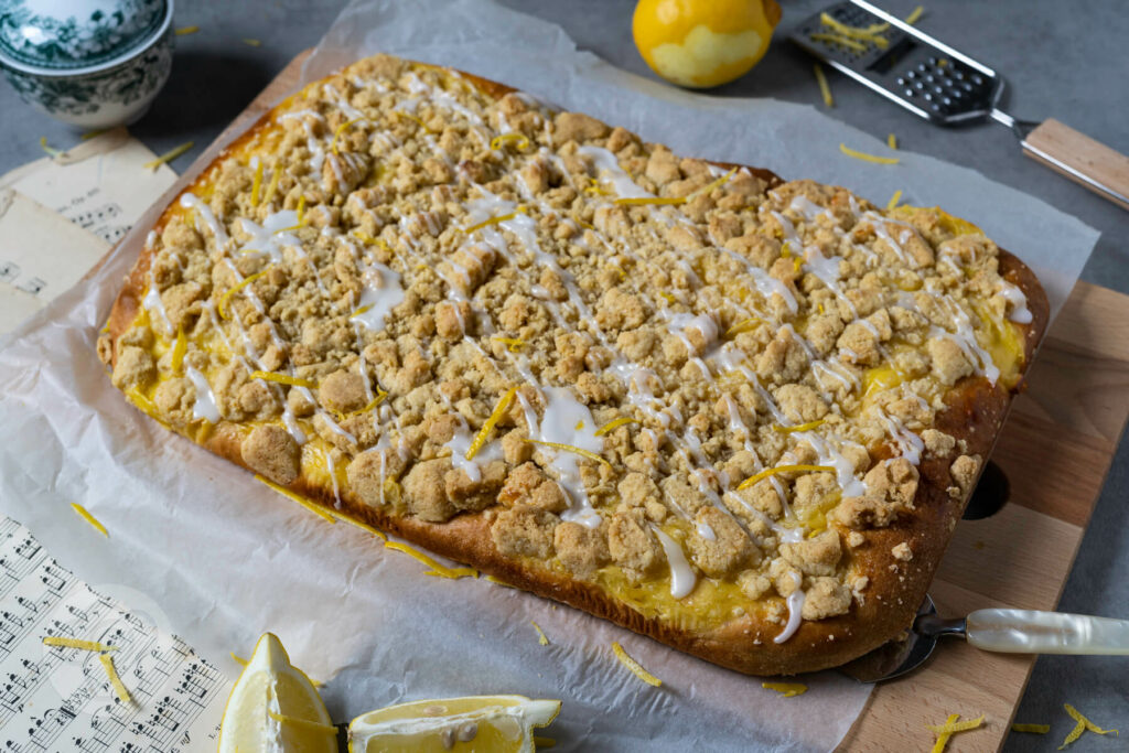 Zitronen Streuselkuchen mit einer Kuchenschaufel. Geschirr und Zitronen im Hintergrund.
