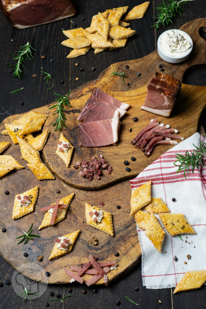 Cracker mit Schwarzwälder Schinken auf einem Brett. Dazu Wacholderbeeren, Schinkenstreifen und Rosmarin.