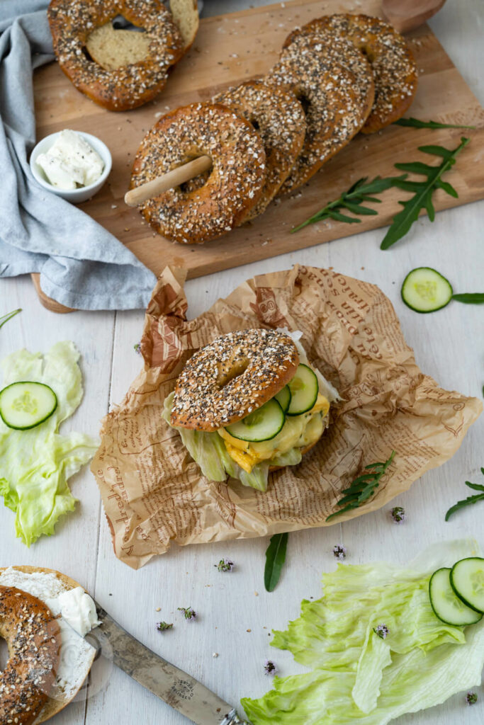 Gebackene Bagel auf einem Kochlöffel aufgereiht, Bagel Sandwich mit Salat, Gurke und Ei. Salat und Gurke drumherum.