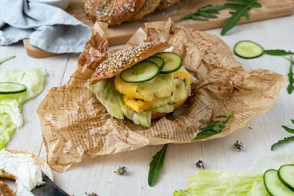 Gebackene Bagel auf einem Kochlöffel aufgereiht, Bagel Sandwich mit Salat, Gurke und Ei. Salat und Gurke drumherum.