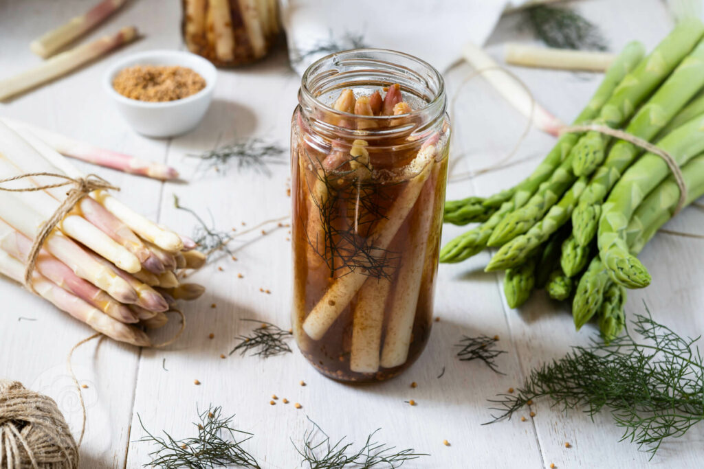 Spargelspitzen im Einmachglas. Im Hintergrund grüner Spargel, Bronzefenchel und Senfkörner.