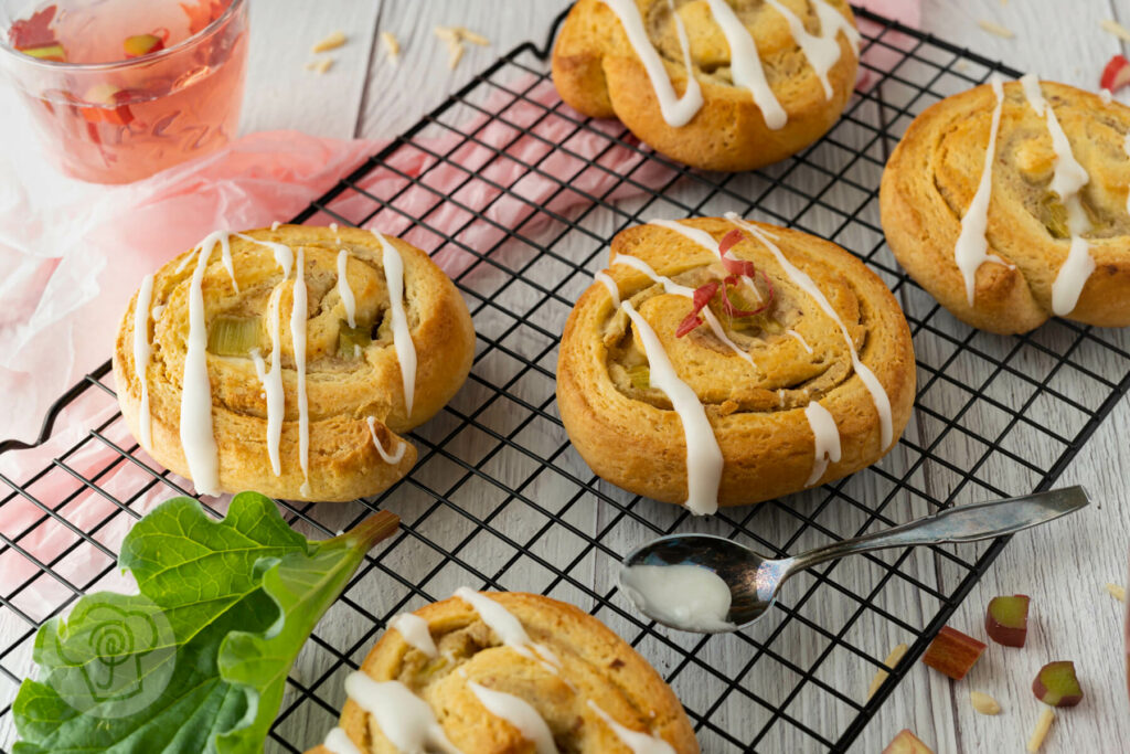 Hefeteigschnecken mit Rhabarber, Marzipan und Zuckerguss auf einem Rost. Im Hintergrund Rhabarberlimonade.