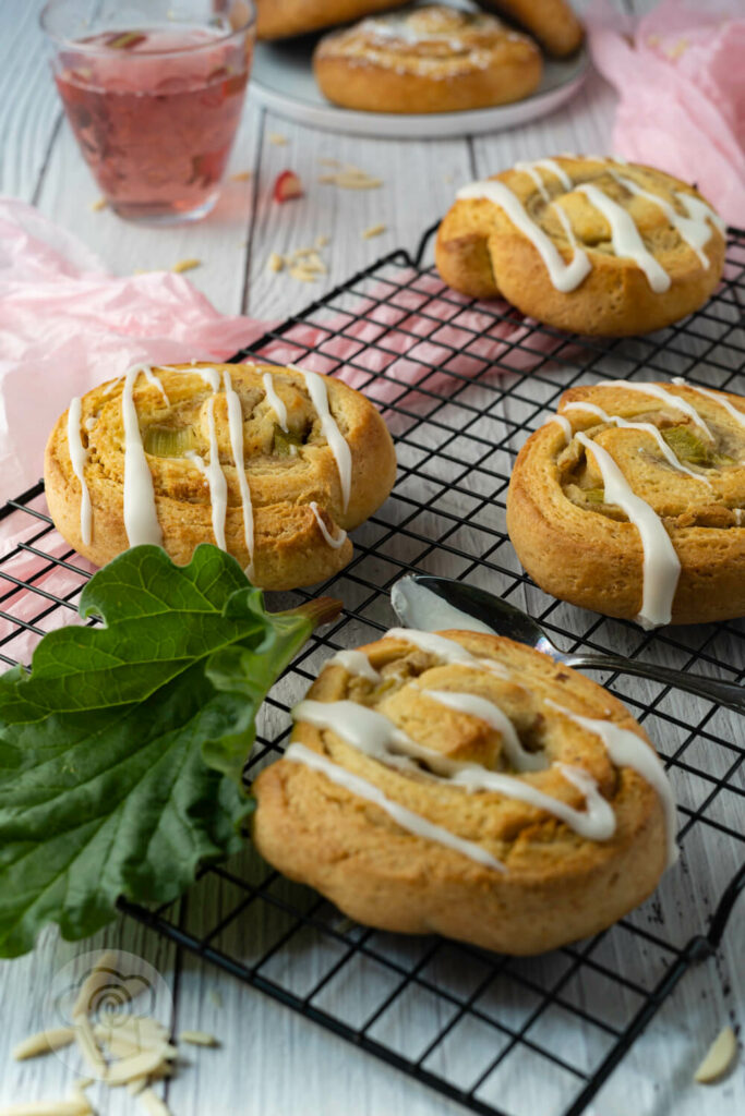 Hefeschnecken mit Rhabarber, Marzipan und Zuckerguss auf einem Rost. Im Hintergrund Rhabarberlimonade.