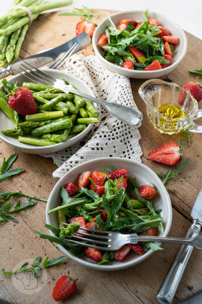 Spargelsalat mit grünem Spargel, Erdbeeren und Rucola in kleinen Tellern.