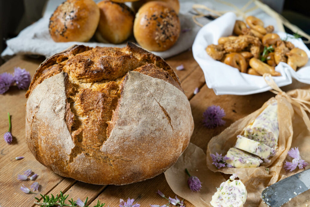 Auffrischbrot für Sauerteigreste auf einem großen Brett, dazu Schnittlauchbutter. Im Hintergrund Brioche Burger Buns und Taralli Pugliesi.