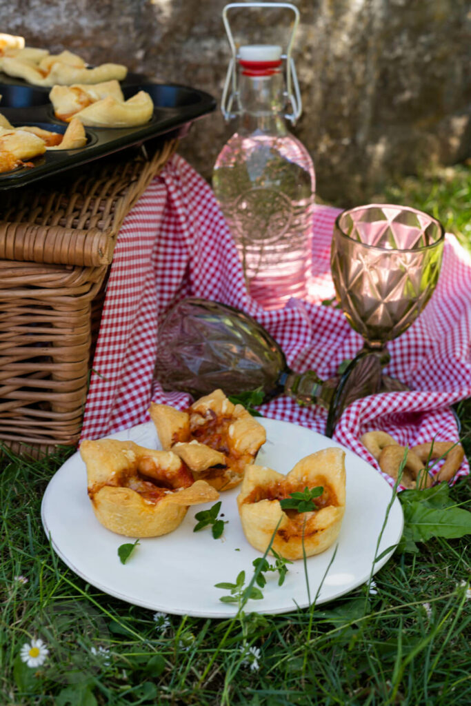 Einfache Pizzamuffins auf einem Teller im Gras. Im Hintergrund Picknickkorb und Gläser