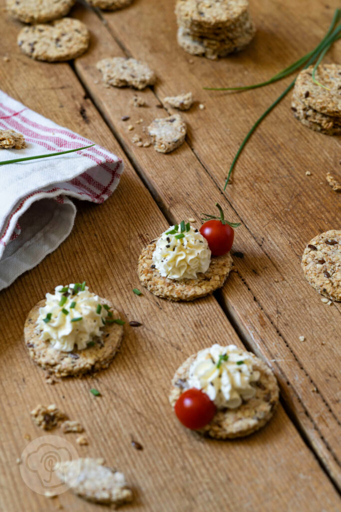 Cracker auf einem Brett. Teilweise mit einer Frischkäsecreme und Tomaten.