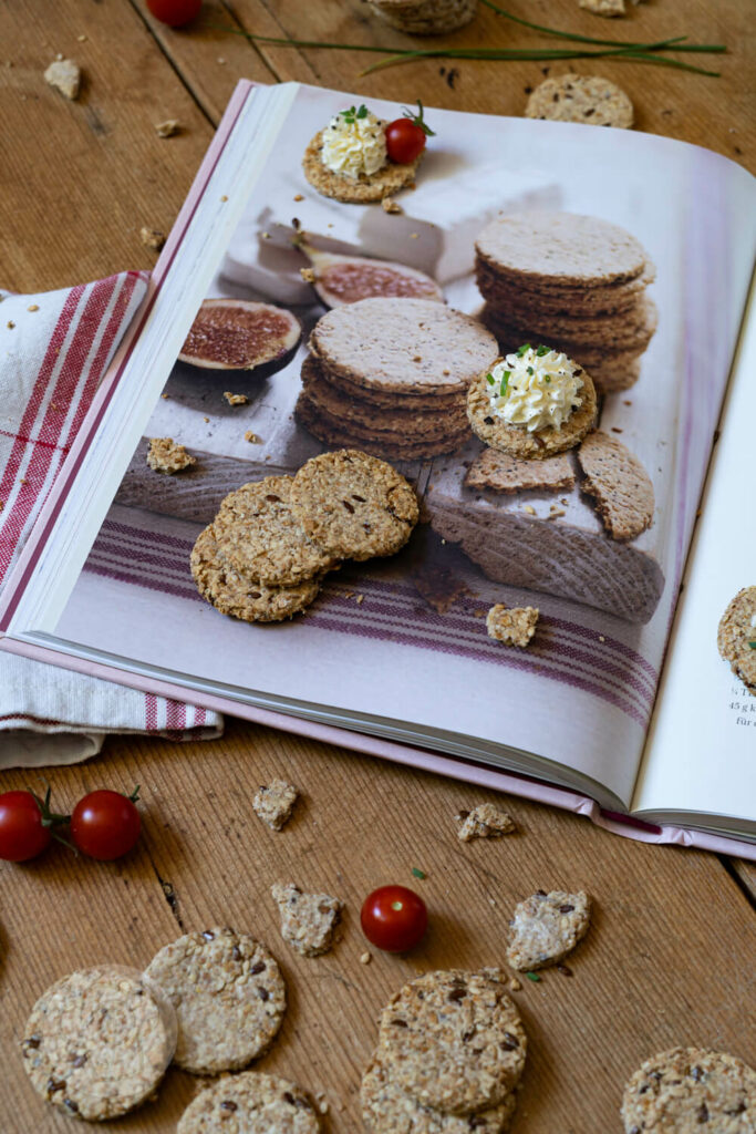 Cracker auf einem Brett. Teilweise mit einer Frischkäsecreme und Tomaten. Aufgeschlagenes Buch.