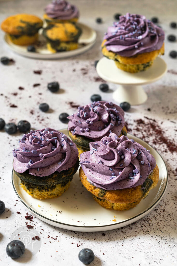 Heidelbeer Cupcakes auf einem Teller im Vordergrund, im Hintergrund ein Teller mit Heidelbeermuffins und einer Etagère