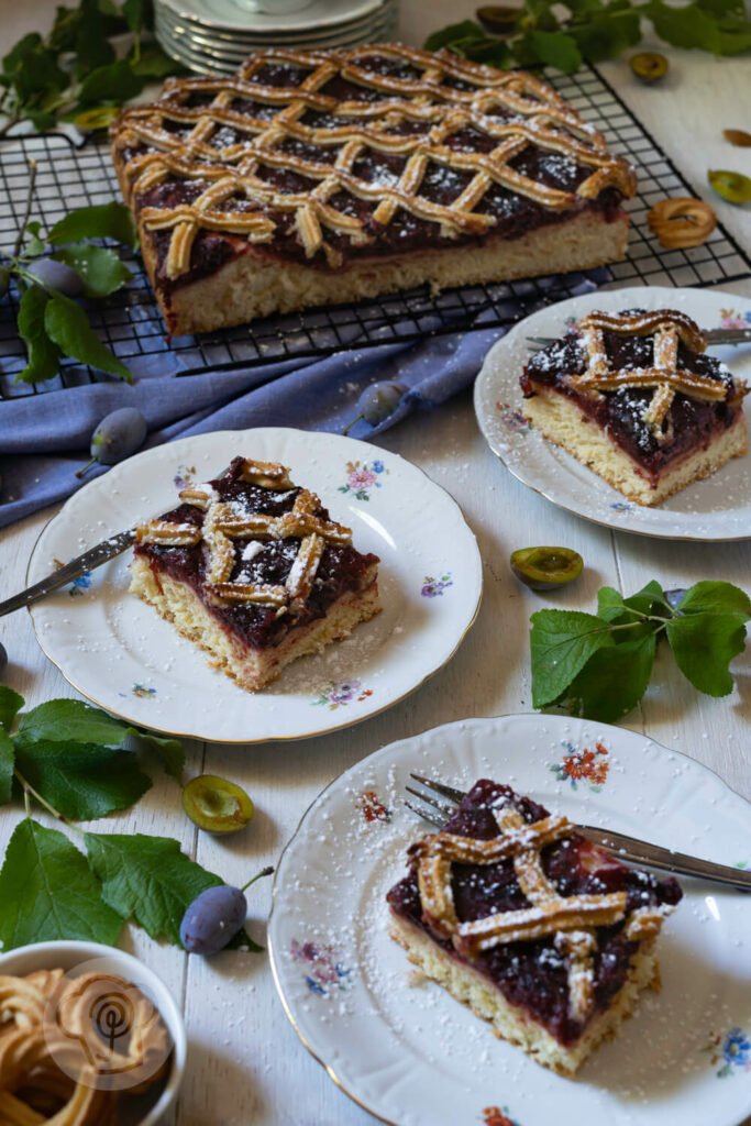 Pflaumenkuchen mit Marzipangitter auf einem Kuchenrost. Im Vordergrund drei Stücke Kuchen auf Kuchentellern verteilt.