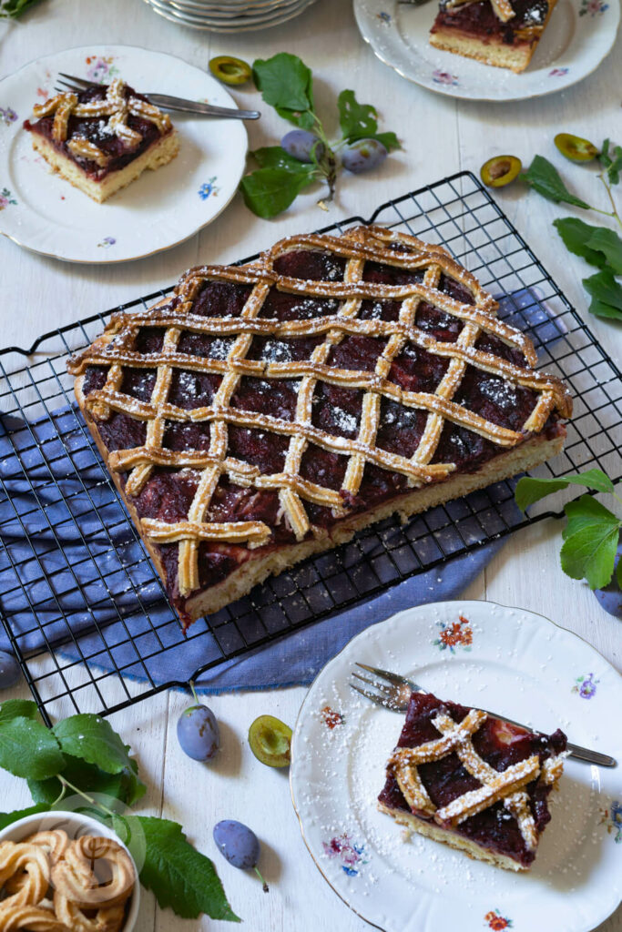 Pflaumenkuchen mit Marzipangitter auf einem Kuchenrost. Kuchenstücke auf einem Tellern.