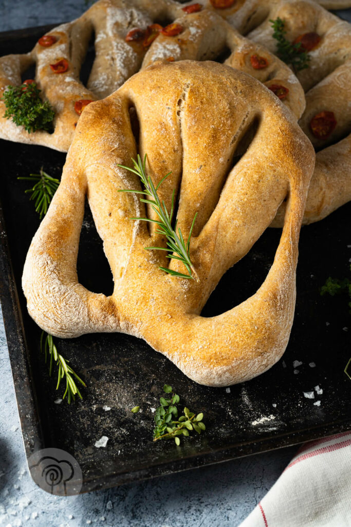 Französische Fougasse auf einem Backblech, mit einem Rosmarinzweig und Tomaten.