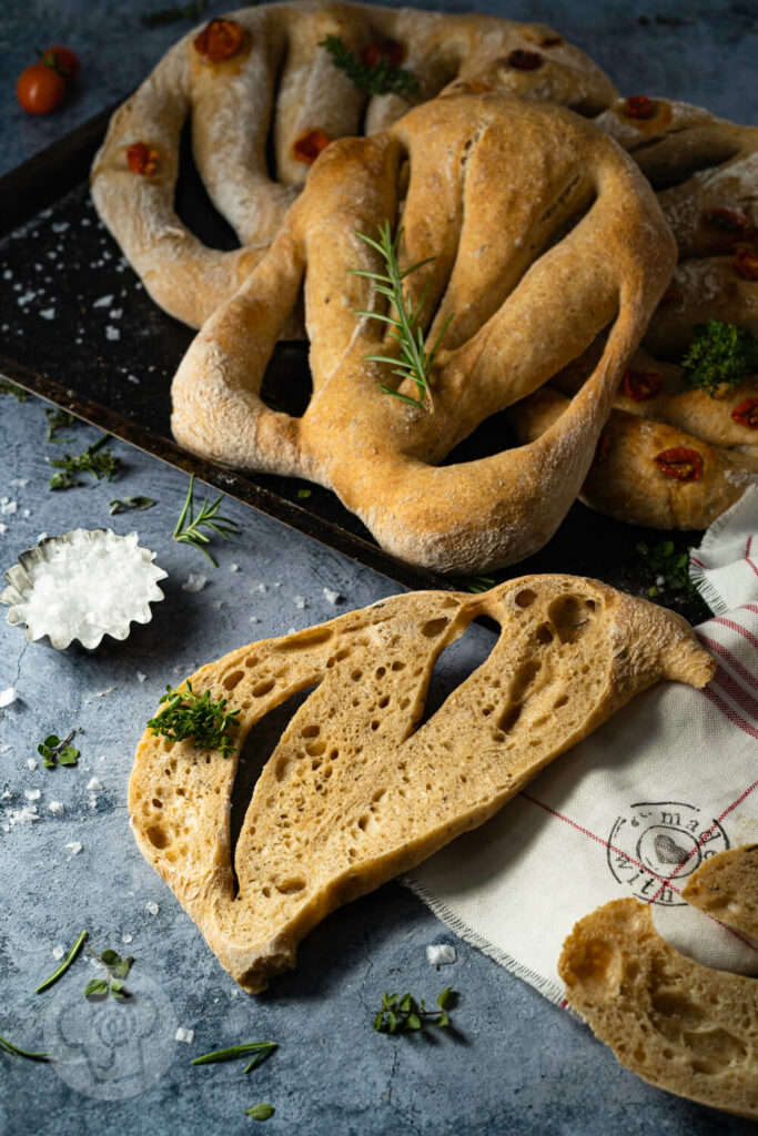 Fougasse mit Kräutern aufgeschnitten, im Hintergrund weitere Hefebrote auf einem Backblech.