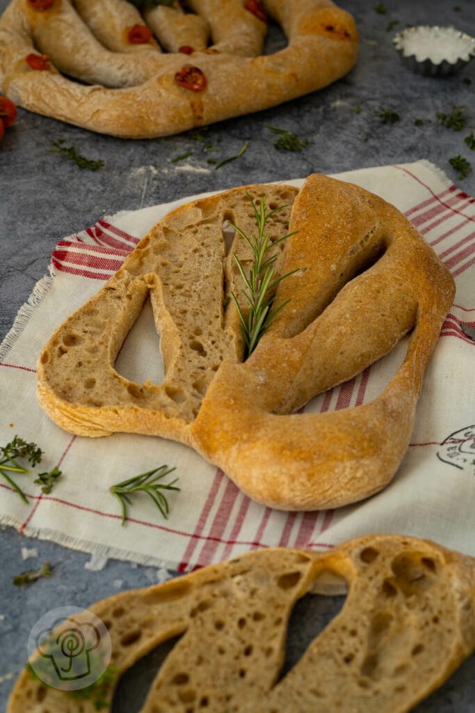 Französische Fougasse zur Hälfte aufgeschnitten auf einem rot-weißen Tuch mit Kräutern.