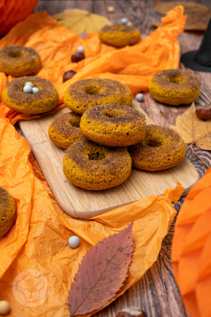 Kürbis Donuts auf einem Holzbrett. Im Hintergrund Hexenhüte, Kastanien und Blätter.