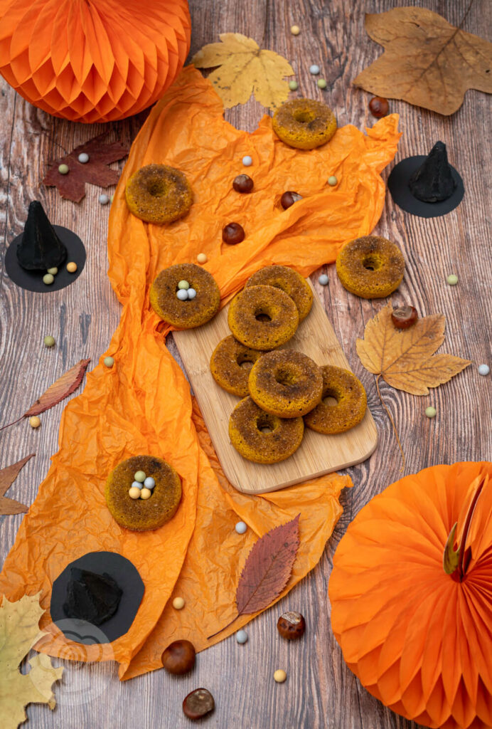 Kürbis Donuts auf einem Holzbrett. Im Hintergrund Hexenhüte, Kastanien und Blätter. Von oben fotografiert.