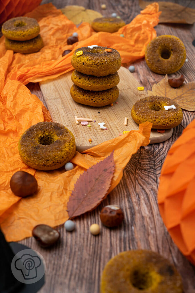 Kürbis Donuts auf einem Holzbrett. Drei Donuts gestapelt. Im Hintergrund Hexenhüte, Kastanien und Blätter.