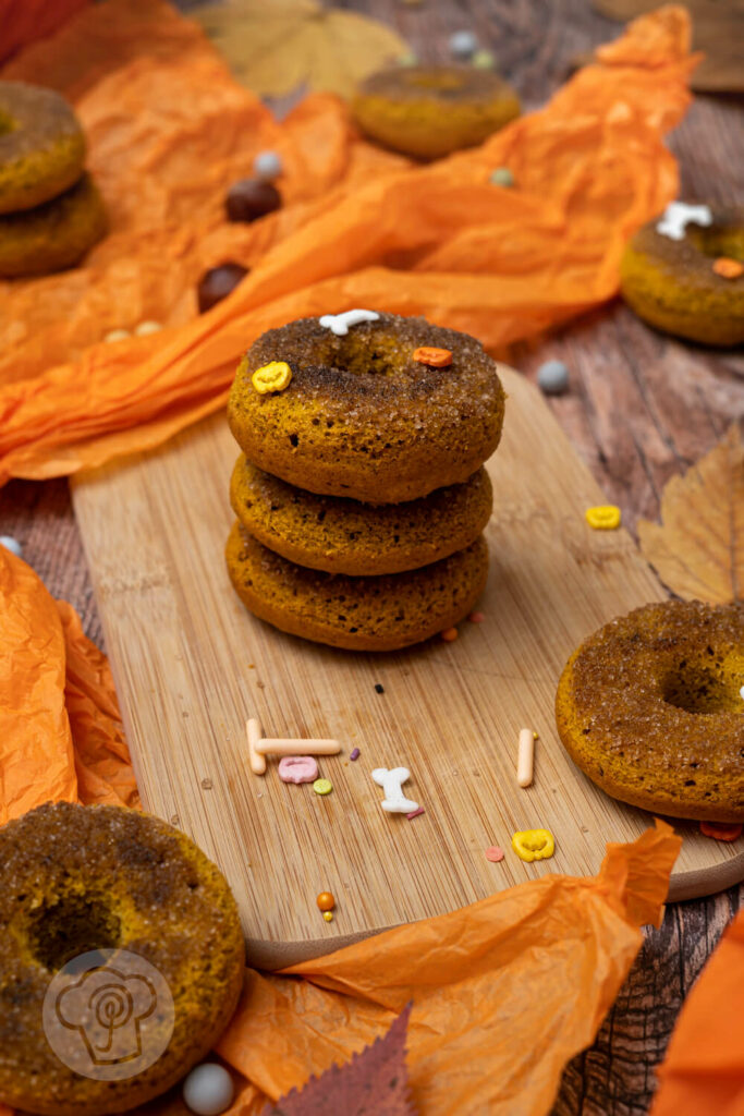 Kürbis Donuts auf einem Holzbrett. Drei Donuts gestapelt. Im Hintergrund Hexenhüte, Kastanien und Blätter.