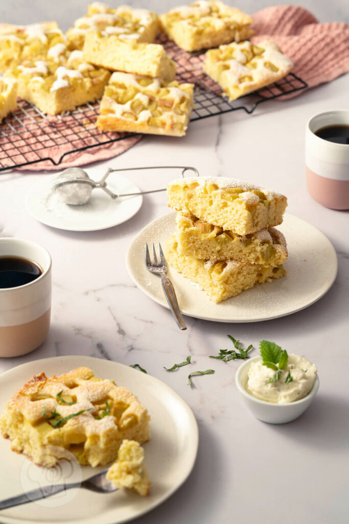 Schneller Rhabarberkuchen vom Blech, in Stücke geschnitten, auf einem Kuchengitter arrangiert und auf einem Kuchenteller gestapelt. Dazu gibt es geschlagene Sahne mit Zitronenmelisse und frischen Kaffee.